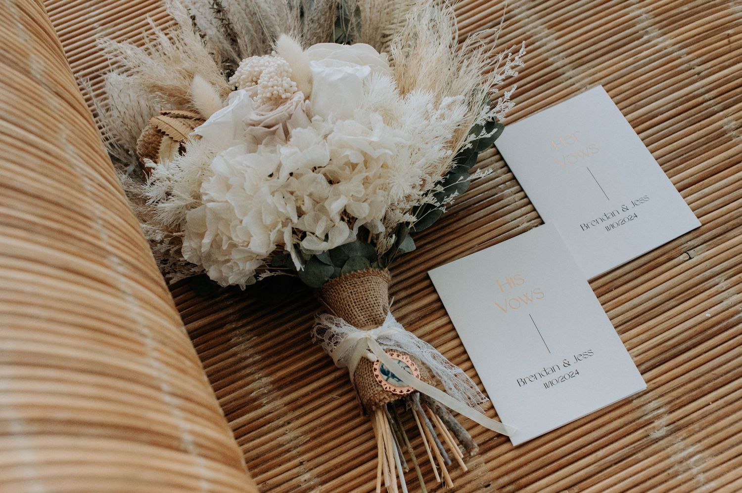 Elegant white floral bouquet with peonies and dried flowers tied with lace ribbon next to invitation cards on wooden surface.