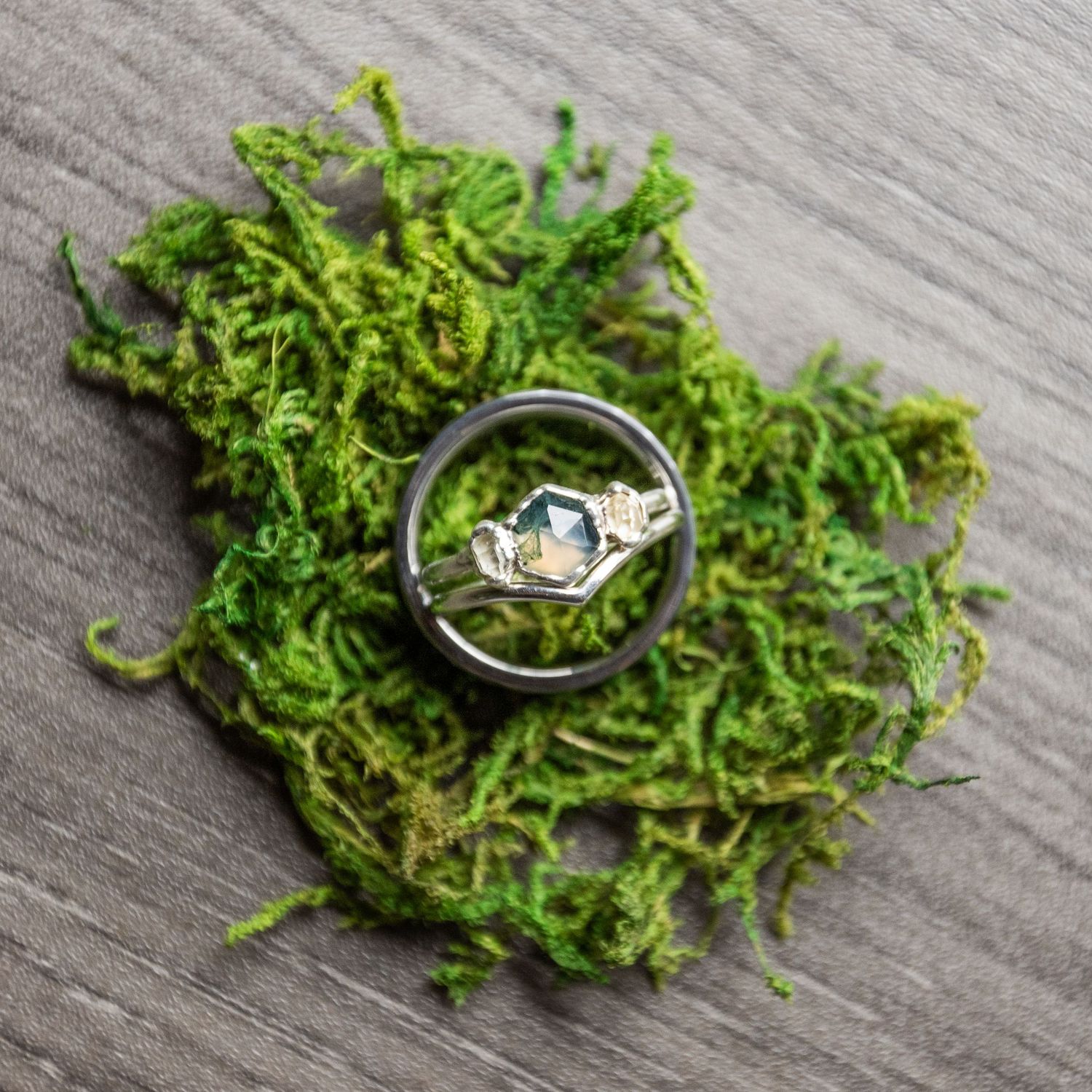 Wedding ring nestled in green moss on gray wooden surface creates rustic engagement photo detail.