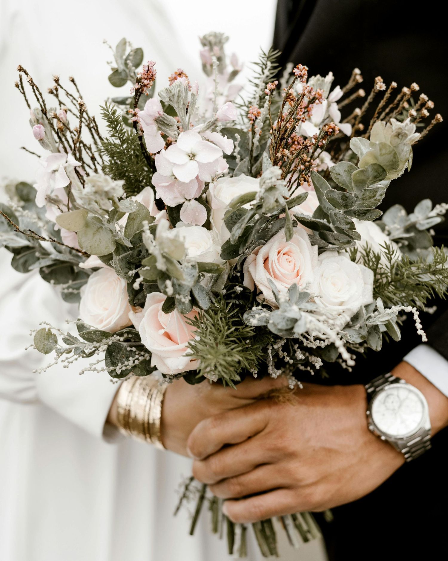 A delicate winter wedding bouquet with blush roses, dusty miller, and dried elements held by hands wearing a silver watch.