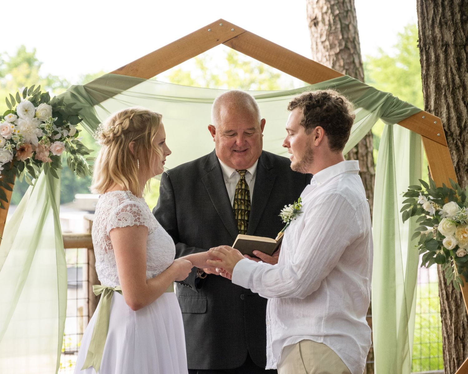 An outdoor wedding ceremony under a wooden arch decorated with floral arrangements and white draping in a natural setting.
