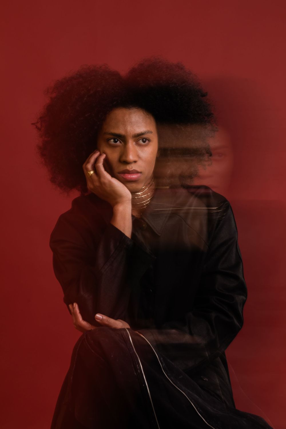 Artistic portrait against red background featuring person with natural afro hair wearing black clothing in moody lighting.