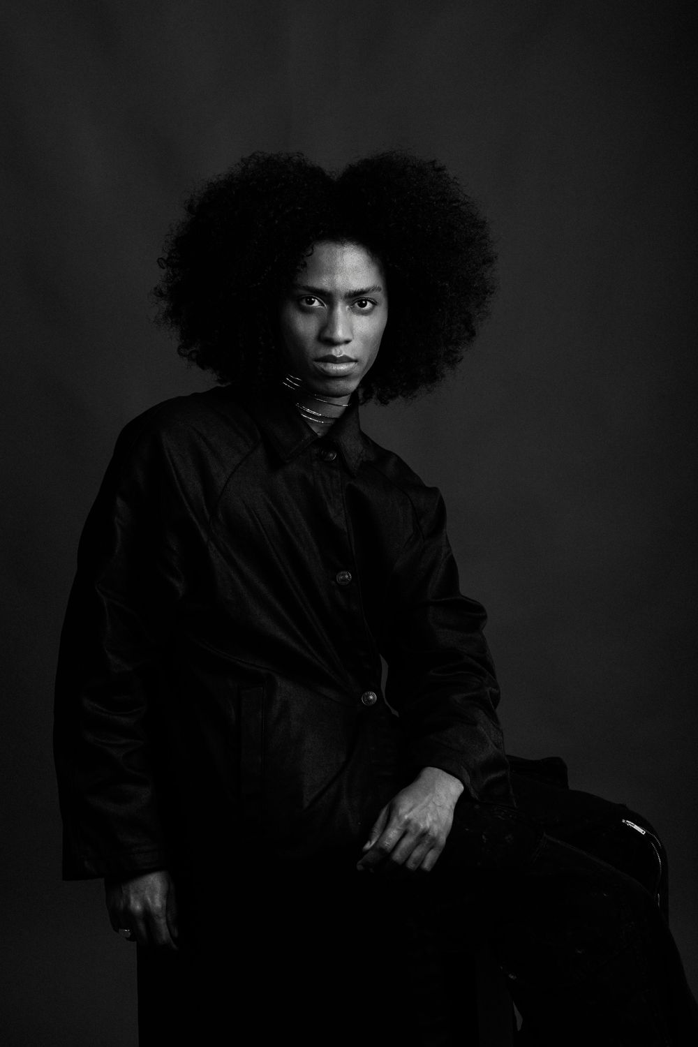 Black and white portrait of a person with natural afro hair wearing a dark coat in dramatic lighting against dark background.