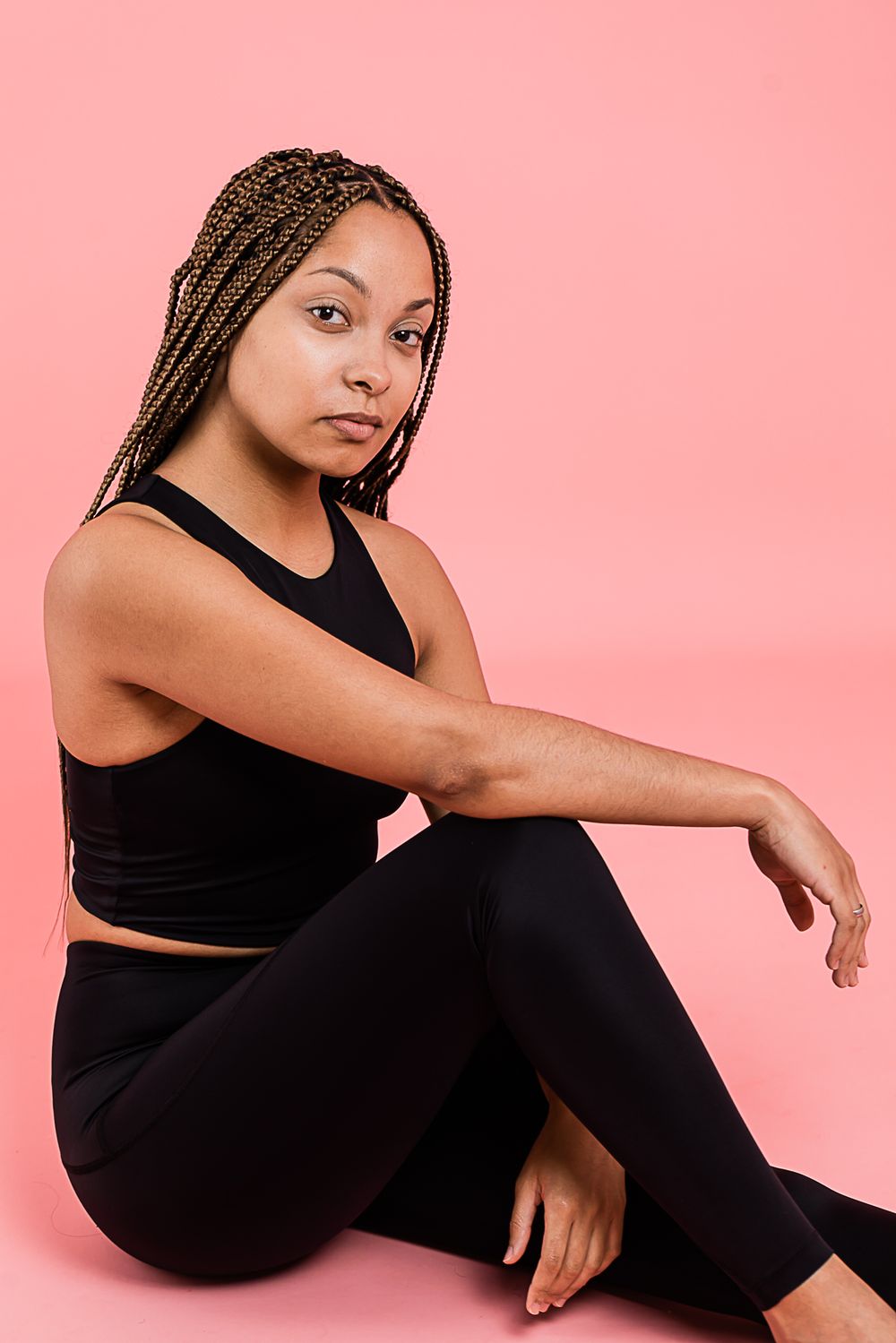Fitness model in black athletic wear sitting gracefully against pink background with braided hair.