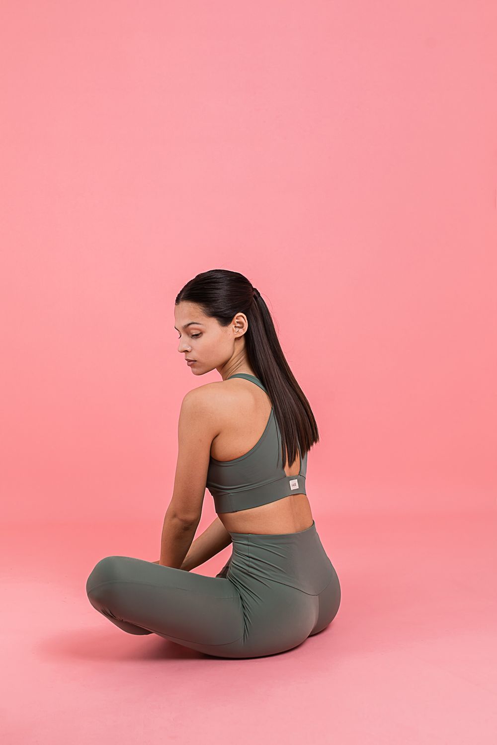 Athletic figure in grey workout outfit sits in yoga pose against pink background during meditation session.