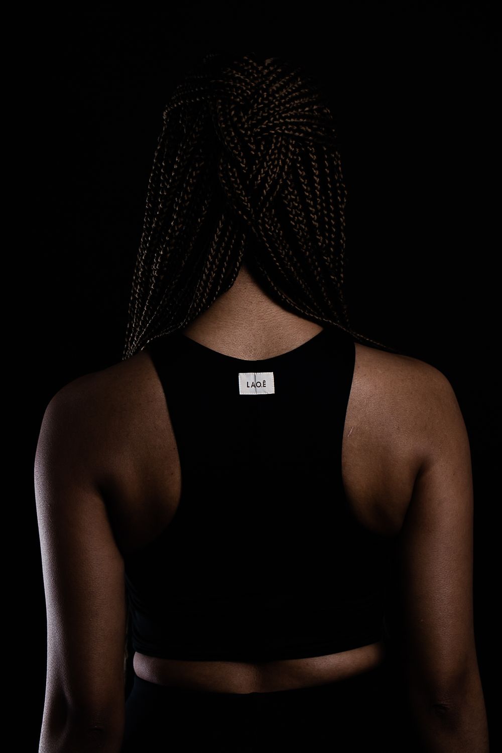 Back view of a person with braided hair wearing a black athletic tank top against a dark background.