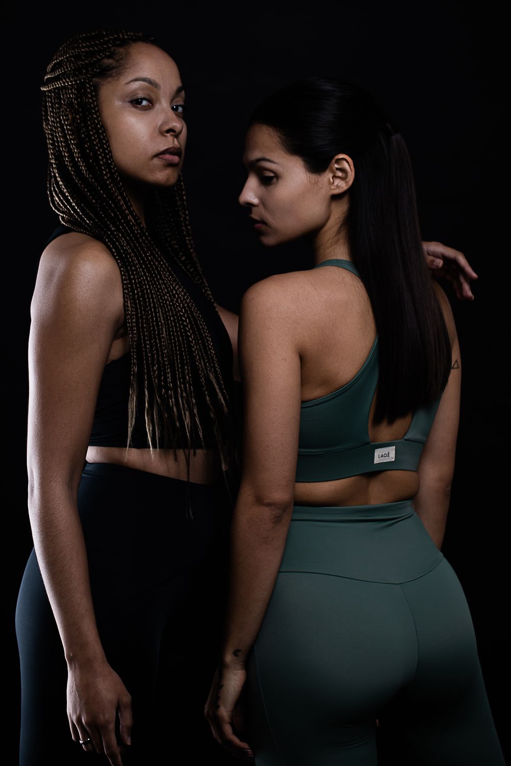 Two athletic women in workout attire pose together against dark background in dramatic lighting.