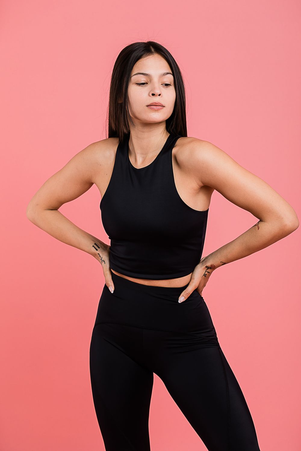 Athletic figure in black workout attire with hands on hips poses confidently against pink background.