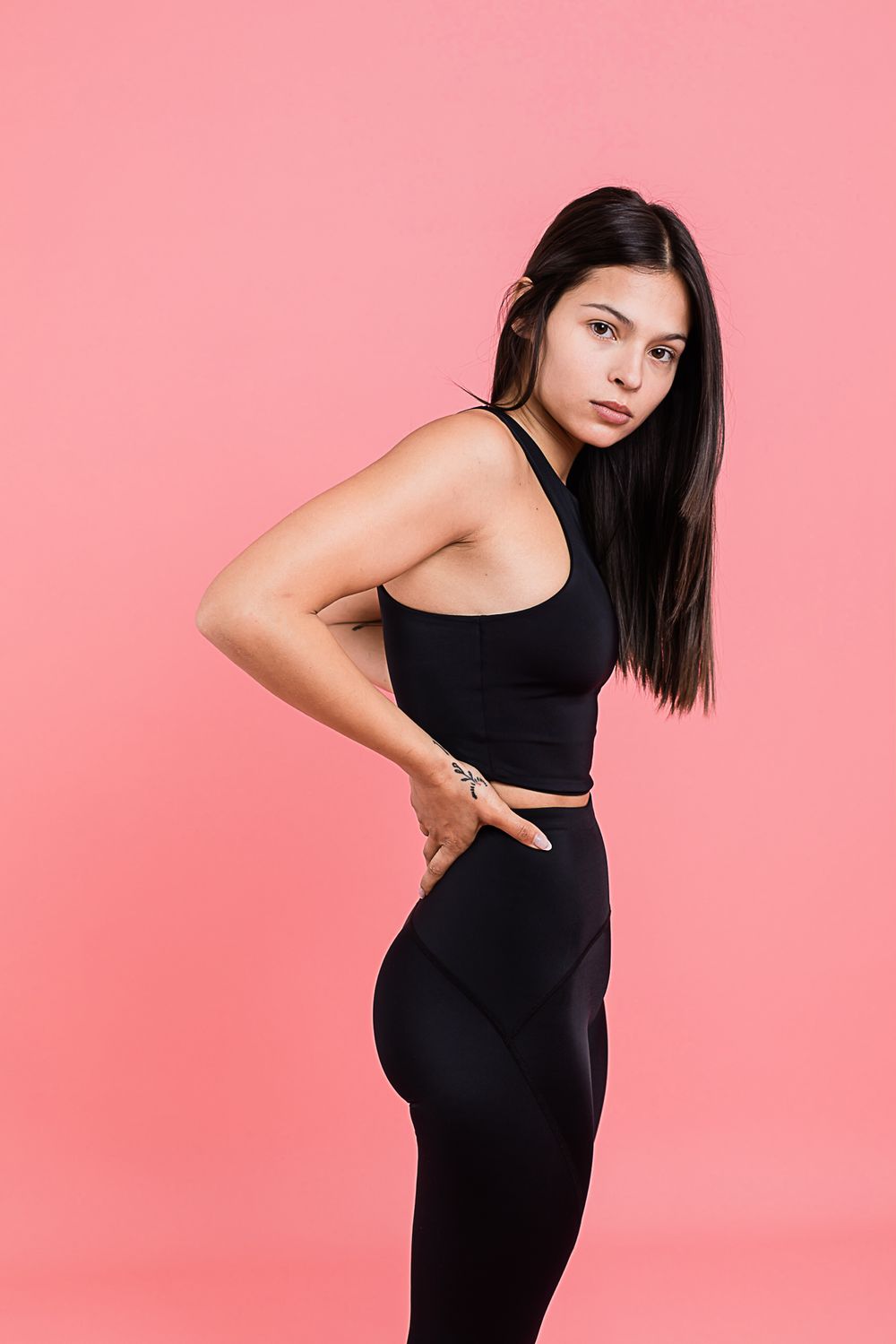 Athletic woman in black workout attire posing against pink background showing fitness form and posture.