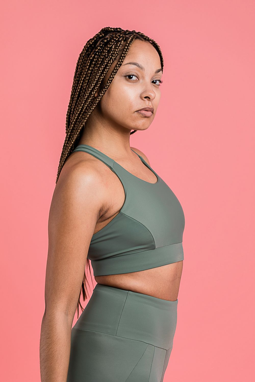 Athletic model wearing gray sports bra and leggings poses against pink backdrop with braided hair and neutral expression.