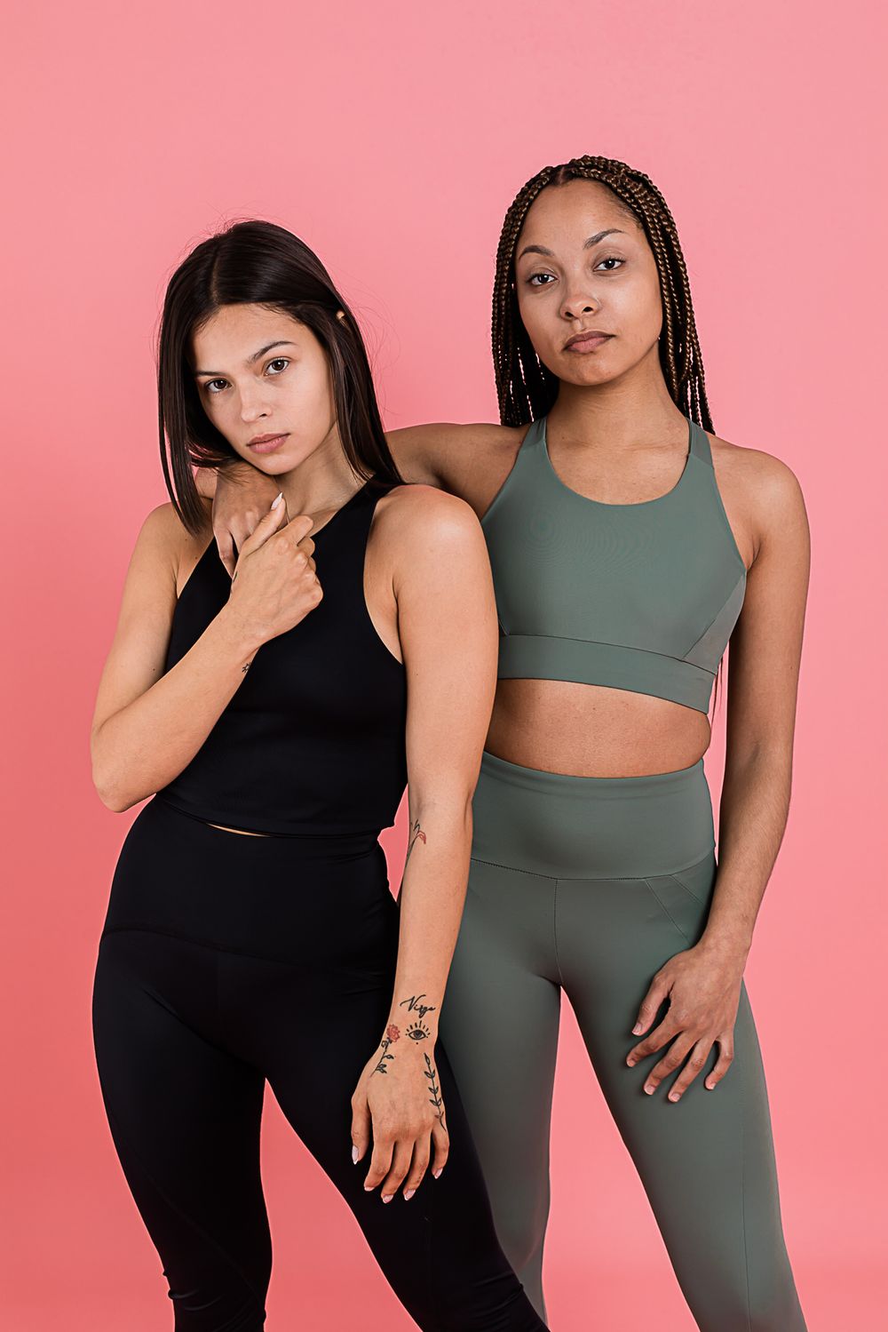 Two fitness models pose in athletic wear featuring a black workout set and a sage green crop top and leggings set against pink.