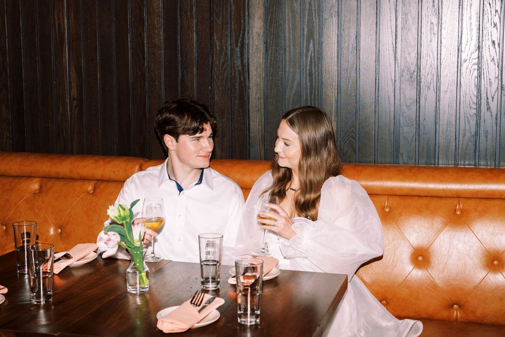 A couple shares conversation and drinks while sitting at a restaurant's leather booth with a small vase of flowers on the table.