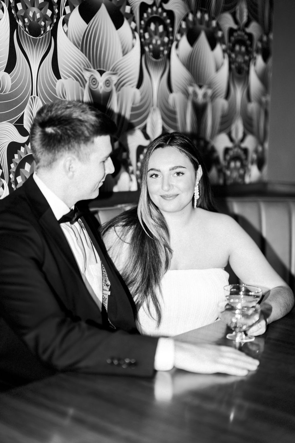 A black and white photo of a couple having drinks at a table with decorative wallpaper in the background.