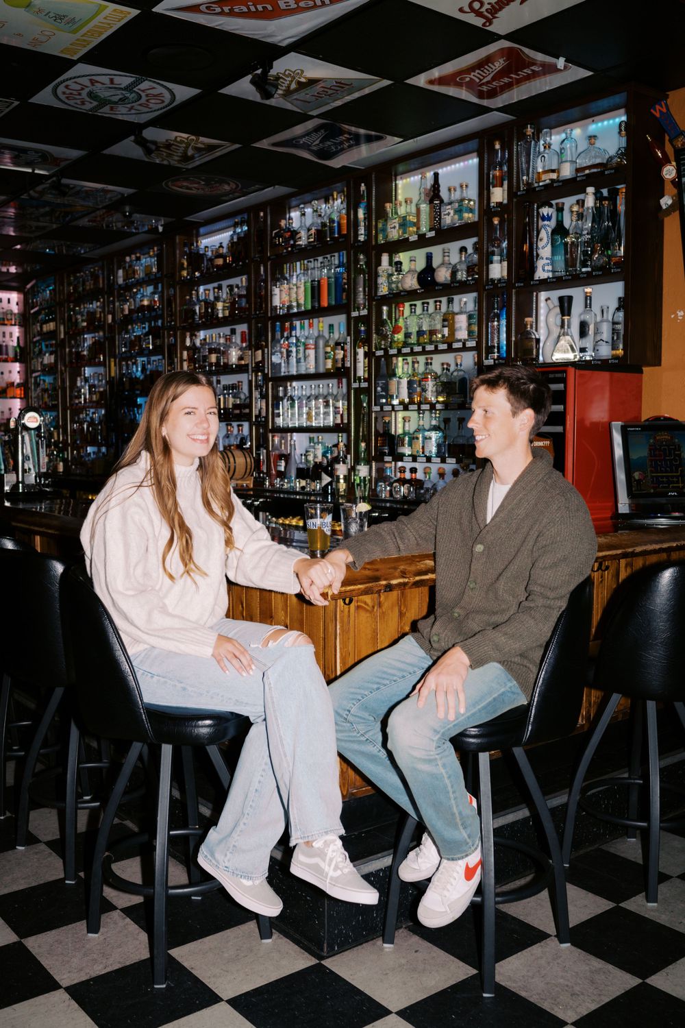 Two people sit and chat at a bar with a large wall display of liquor bottles and checkerboard floor in the background.