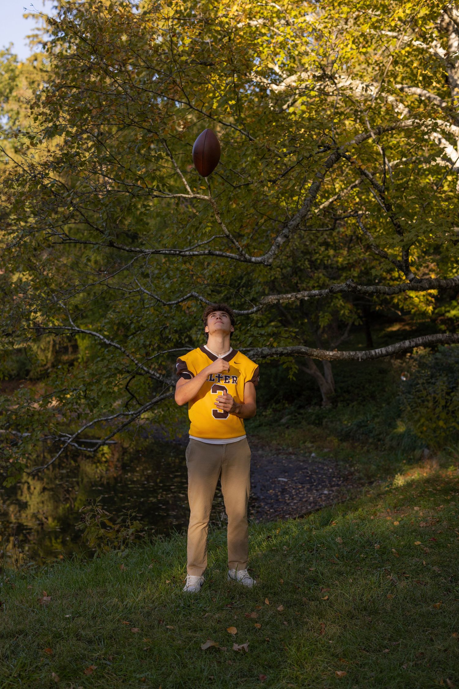 Someone in a yellow top stands on grass while tossing a football in the air on an autumn day.