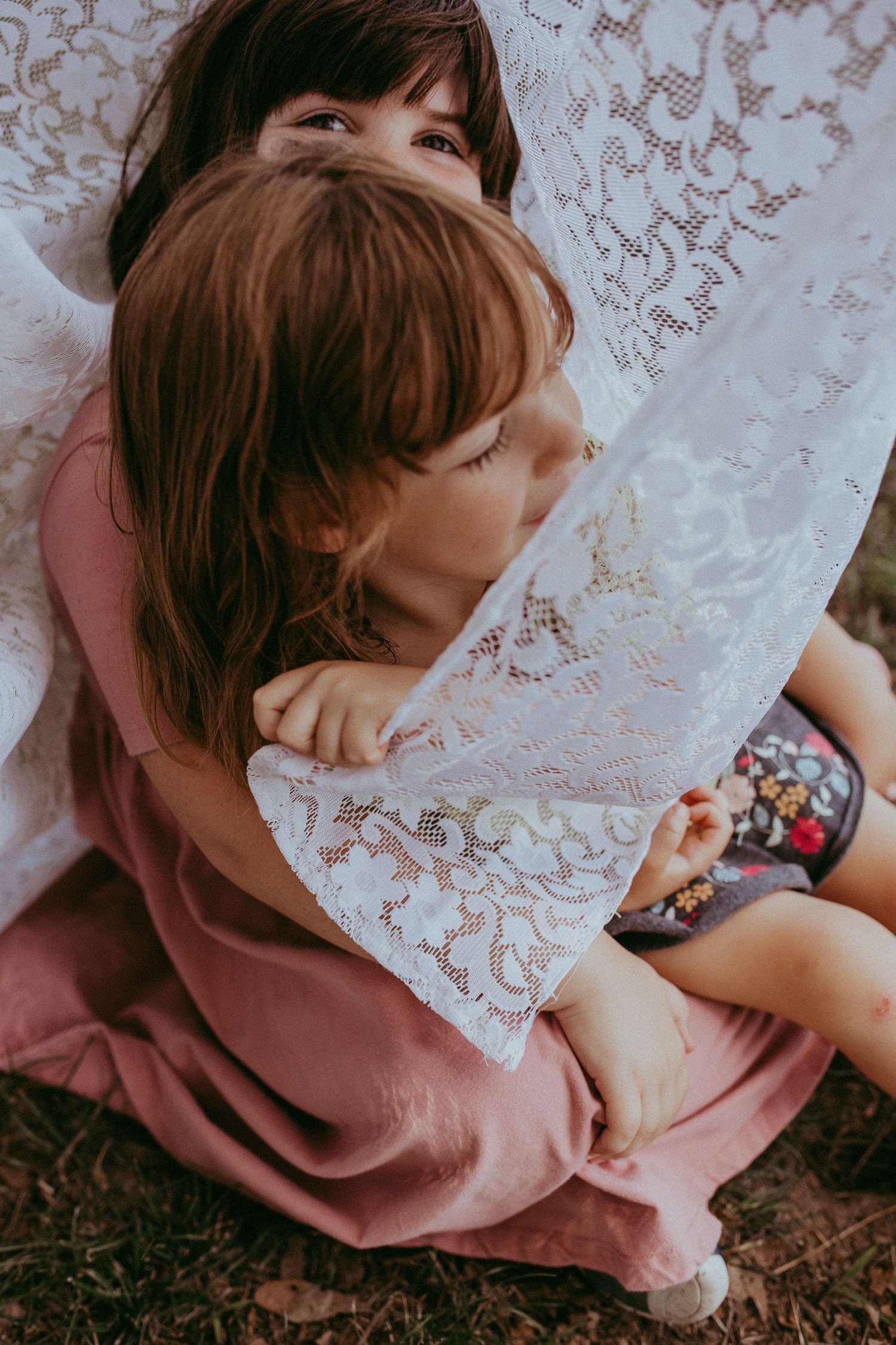 A child in a pink dress plays with a delicate white lace curtain while sitting on the ground. Whitney Popp, Virginia.