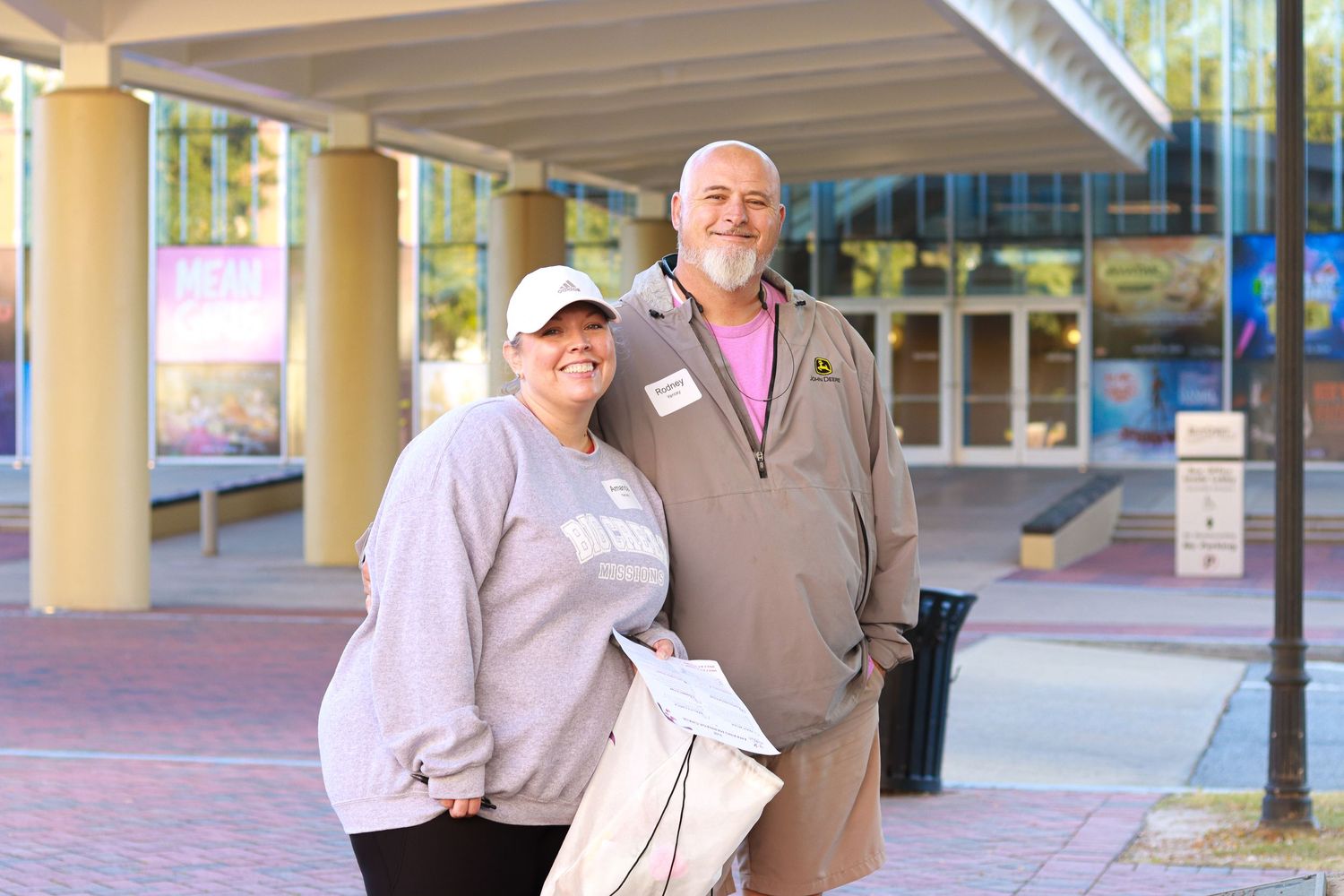 Two people stand together smiling under a covered walkway with columns and brick pavement.
