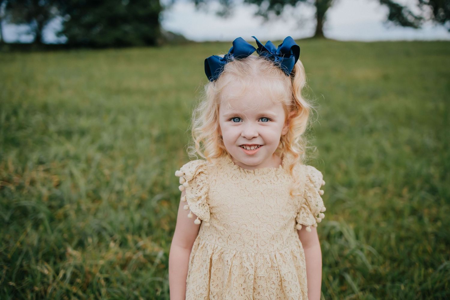Young child in a yellow dress with blue hair bows stands in a grassy outdoor setting on a sunny day.