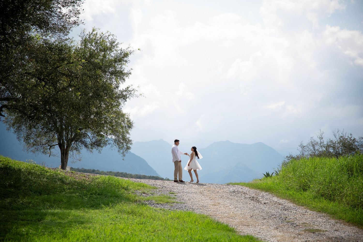 A couple stands on a mountain path surrounded by lush greenery and scenic mountain views in the distance.