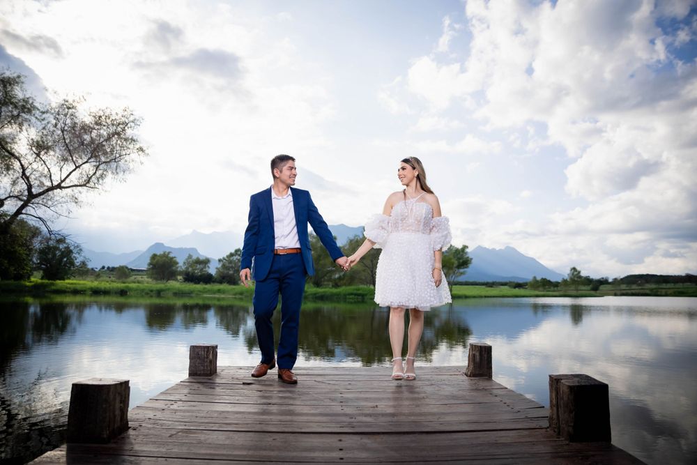 A romantic couple stands on a wooden pier overlooking a serene lake with mountains in the background.