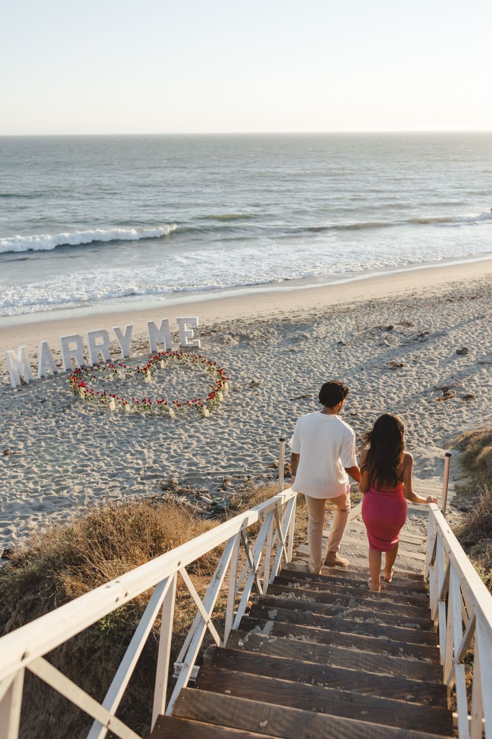 A couple descends white wooden stairs to a sunset beach where 'Marry Me' is written in candles on the sand.