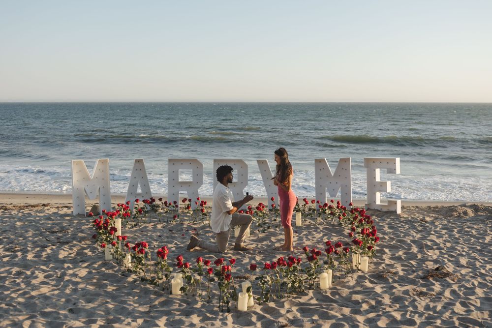 A romantic beach proposal setup with 'MARRY ME' letters and heart-shaped flower arrangement with candles on the sand at sunset.