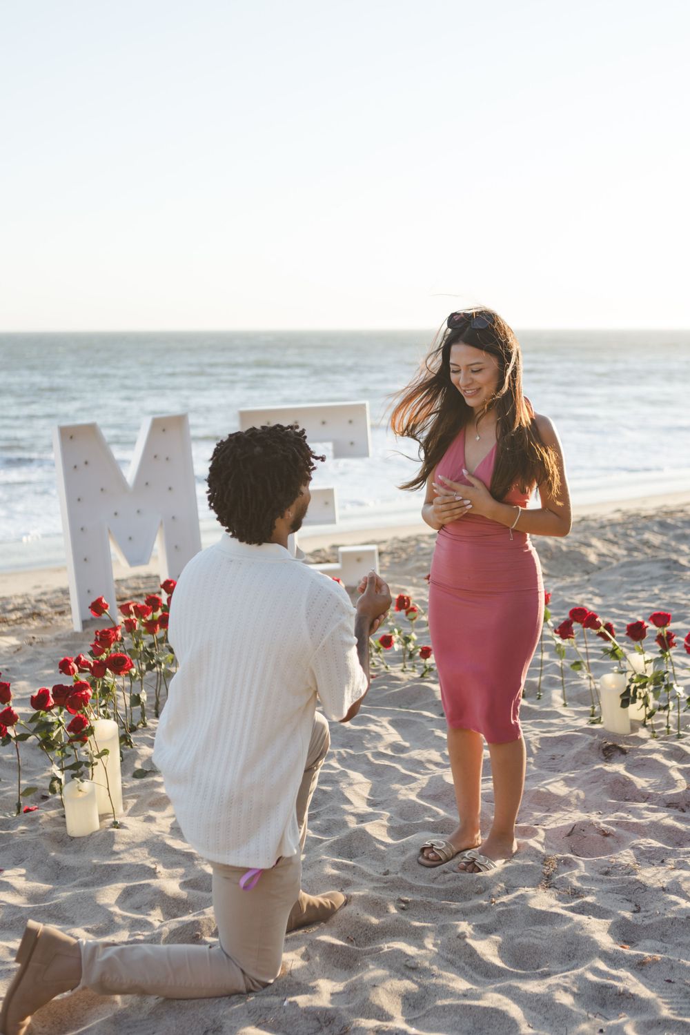 A romantic beach proposal setup with a large letter M decoration, candles and red roses arranged on the sand at sunset.