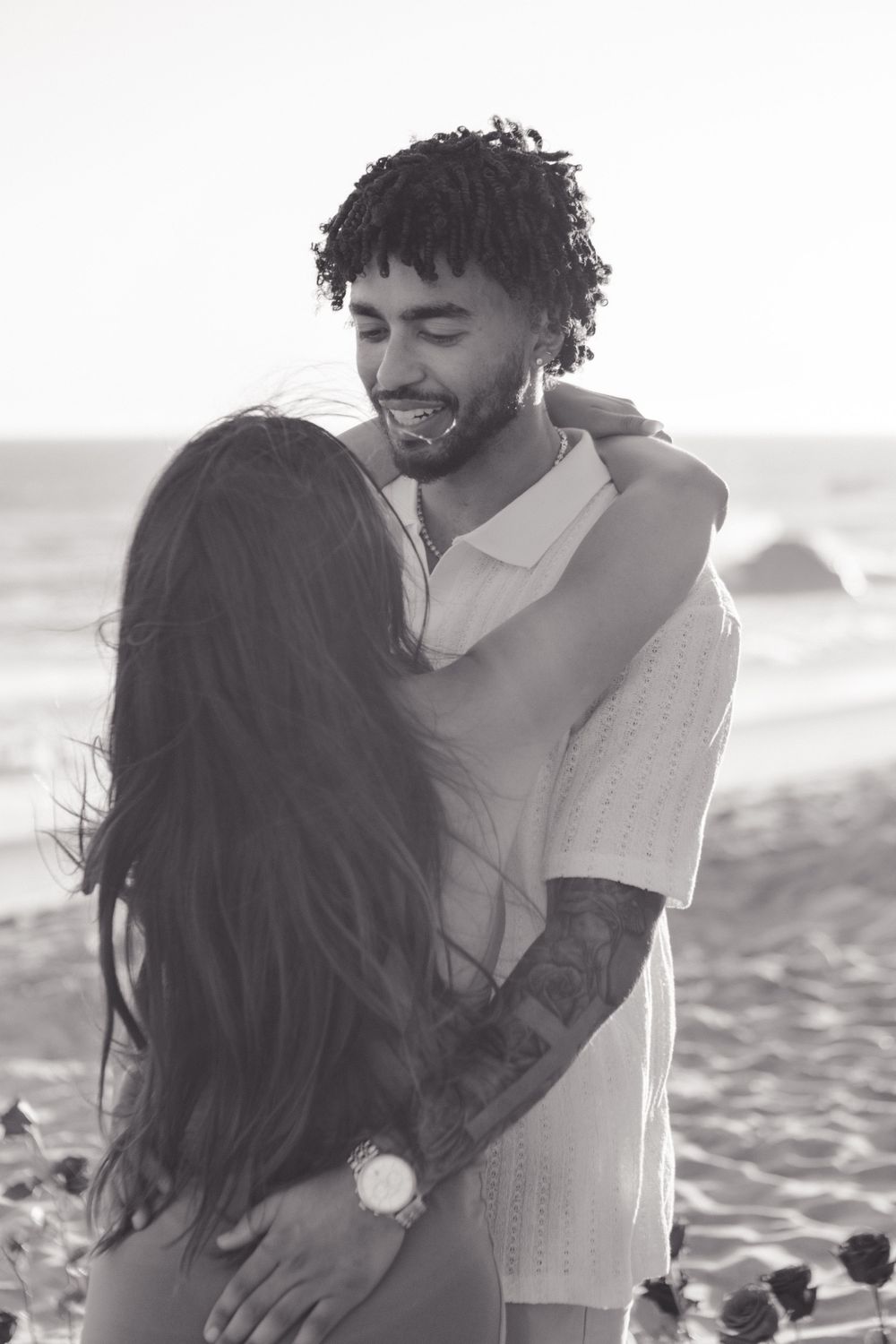 A romantic black and white portrait of a couple embracing and laughing together on a beach at sunset.