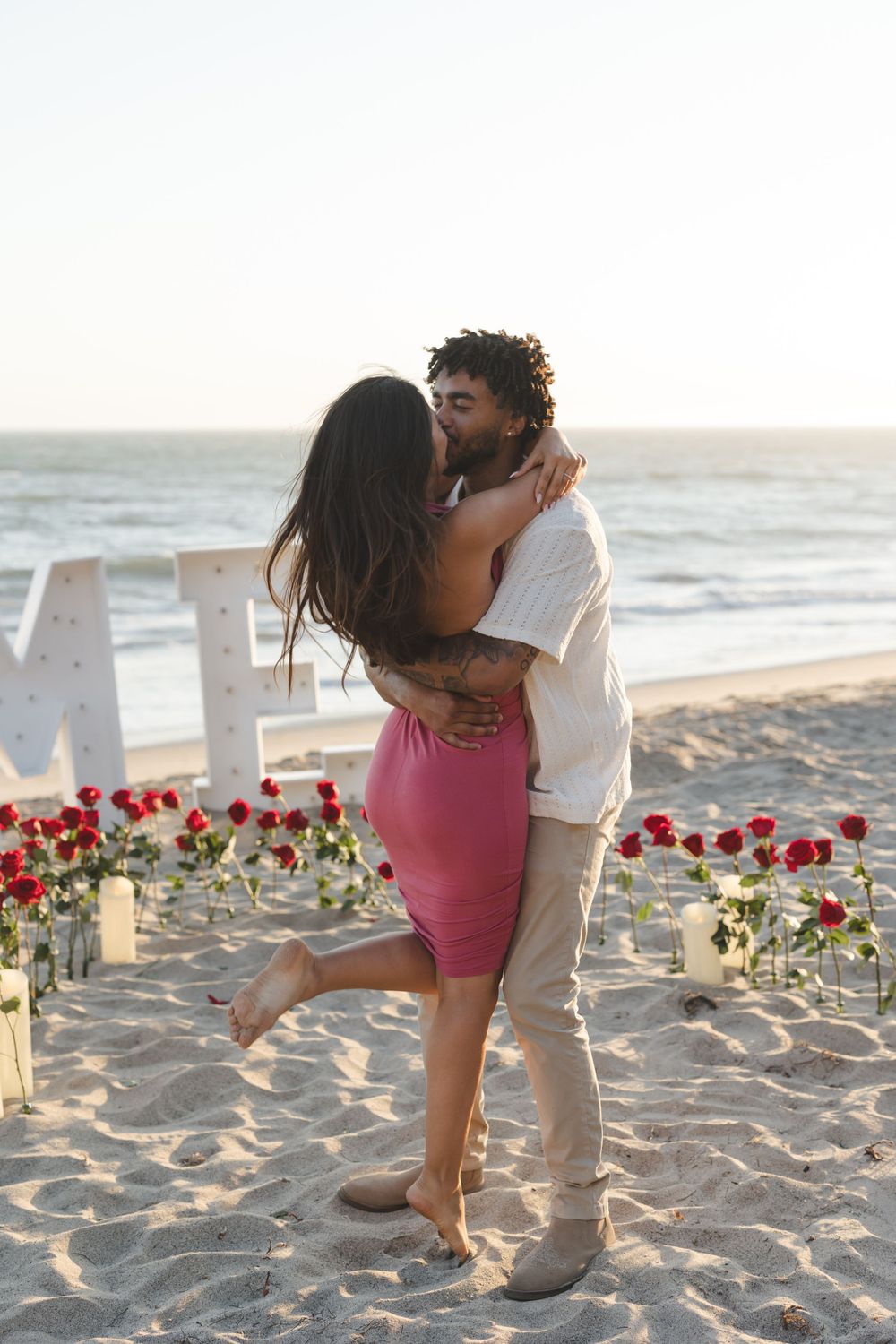 A romantic beach proposal setup with red roses and candles on the sand at sunset along the ocean shore.