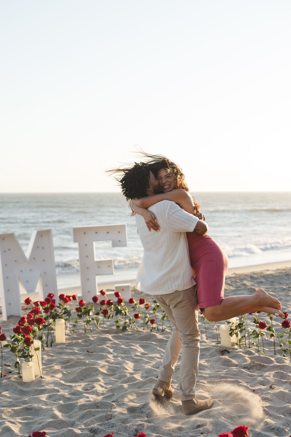 A romantic beach proposal scene with large ME letters and scattered roses on the sand during sunset.
