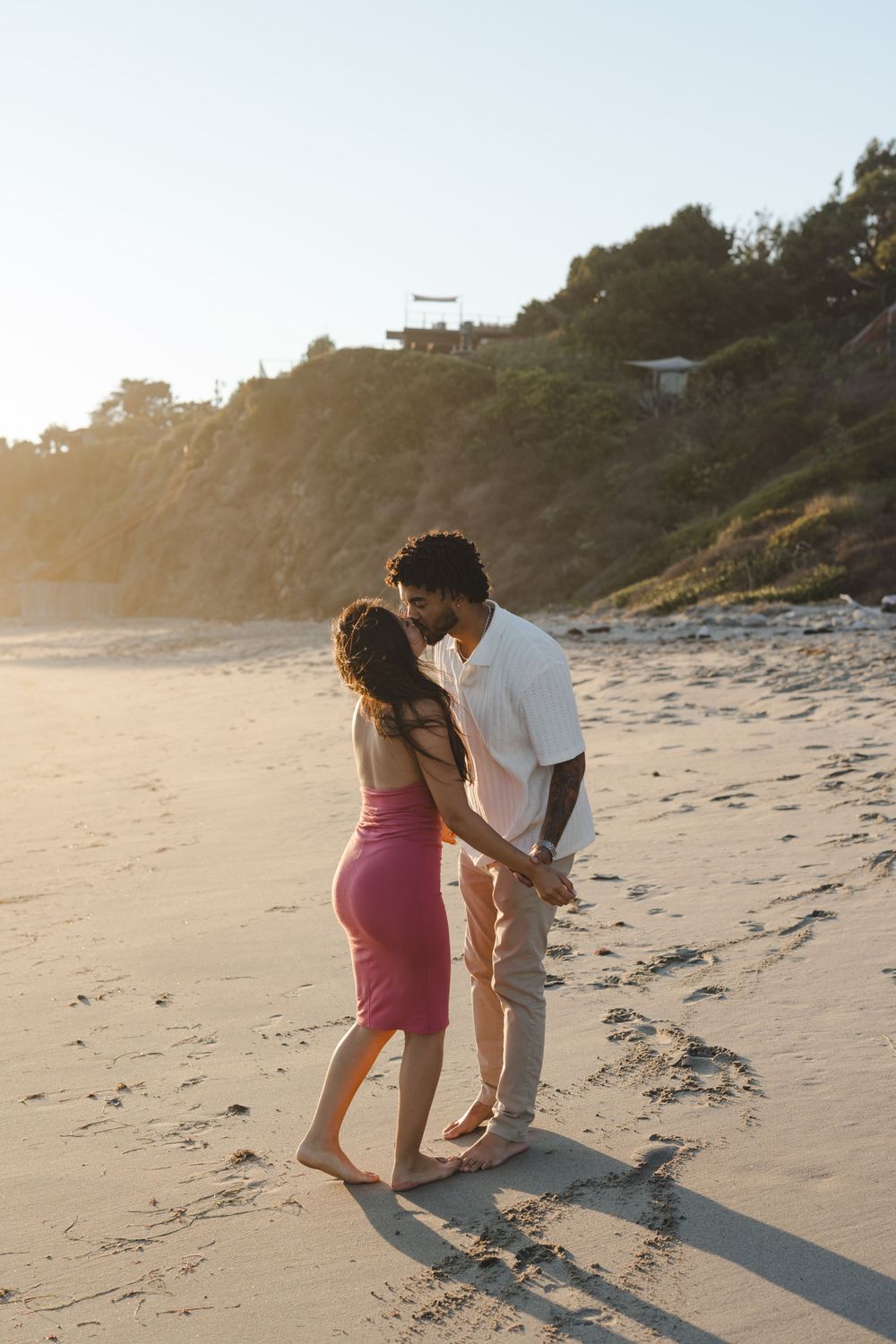 A romantic couple shares an intimate moment on a sunny beach with cliffs and golden sunlight in the background.