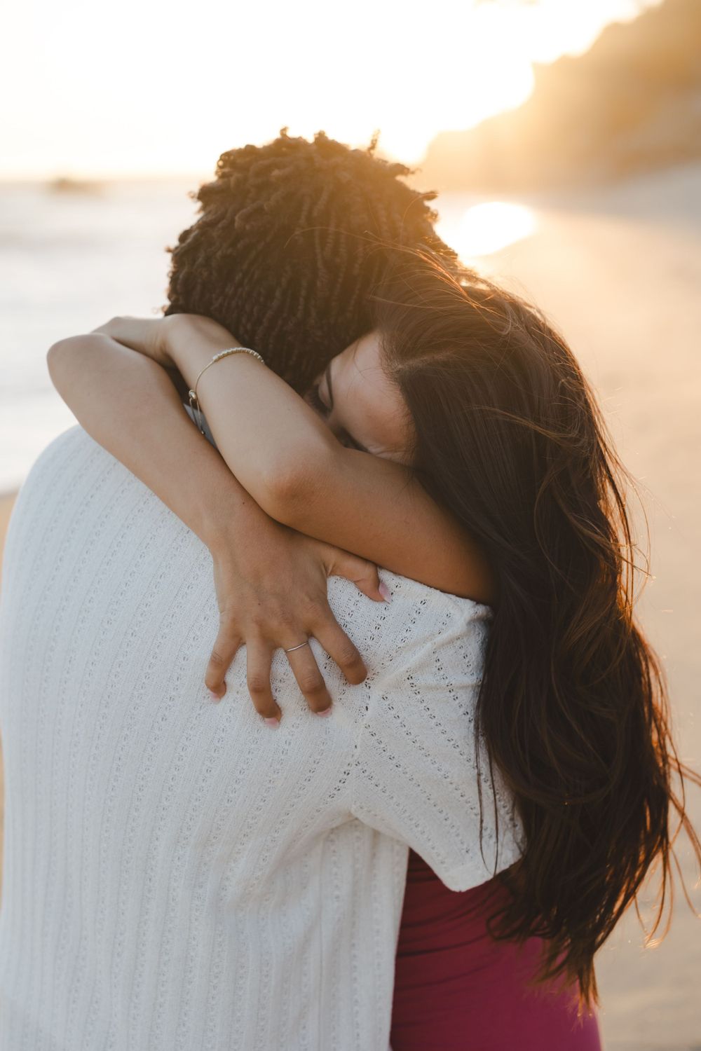 A romantic embrace at sunset between a couple on the beach, one wearing a white sweater.