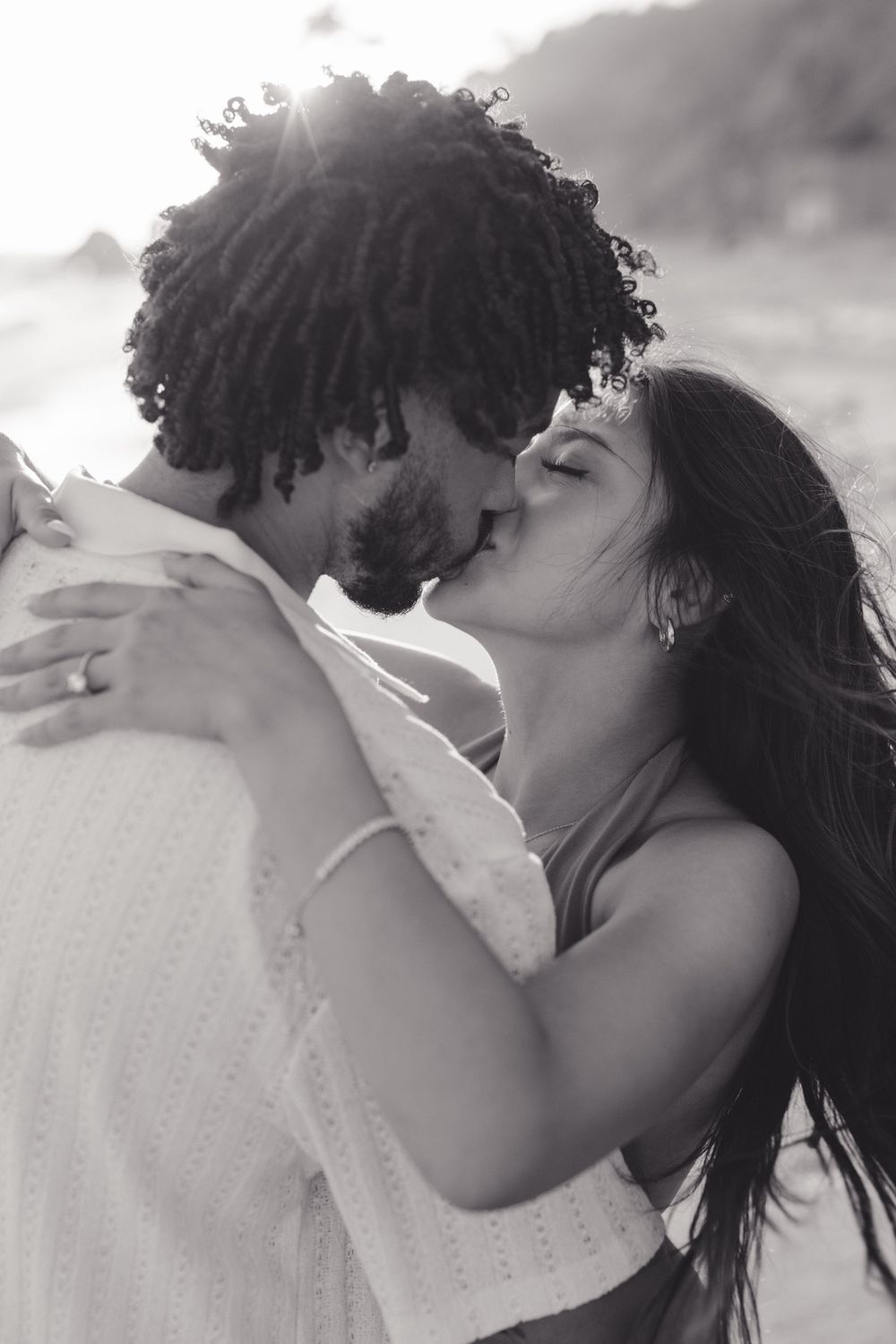 An intimate black and white portrait of a couple embracing on the beach at sunset.