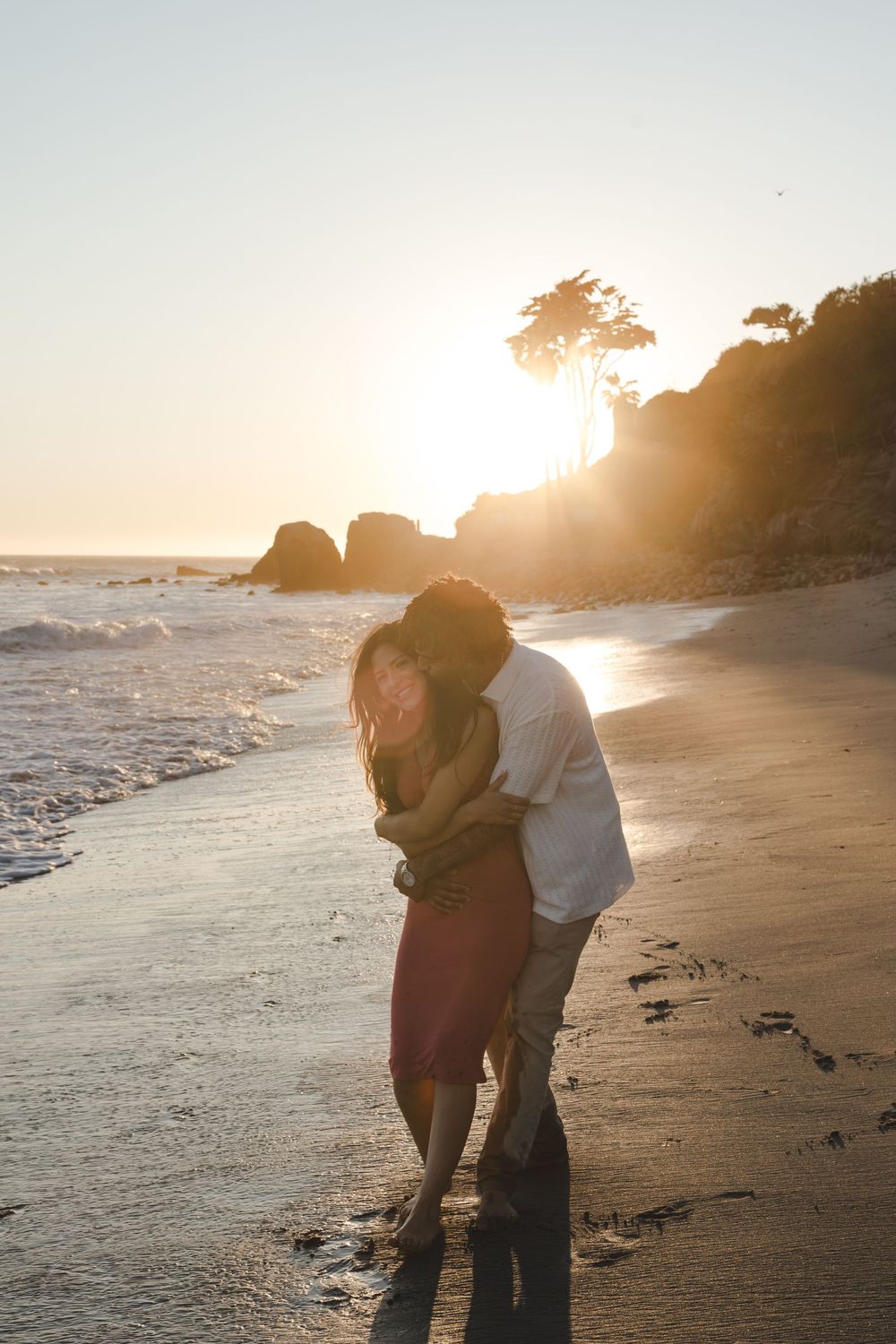 A romantic couple embraces on a beach at sunset with waves and rocks silhouetted in the golden light.