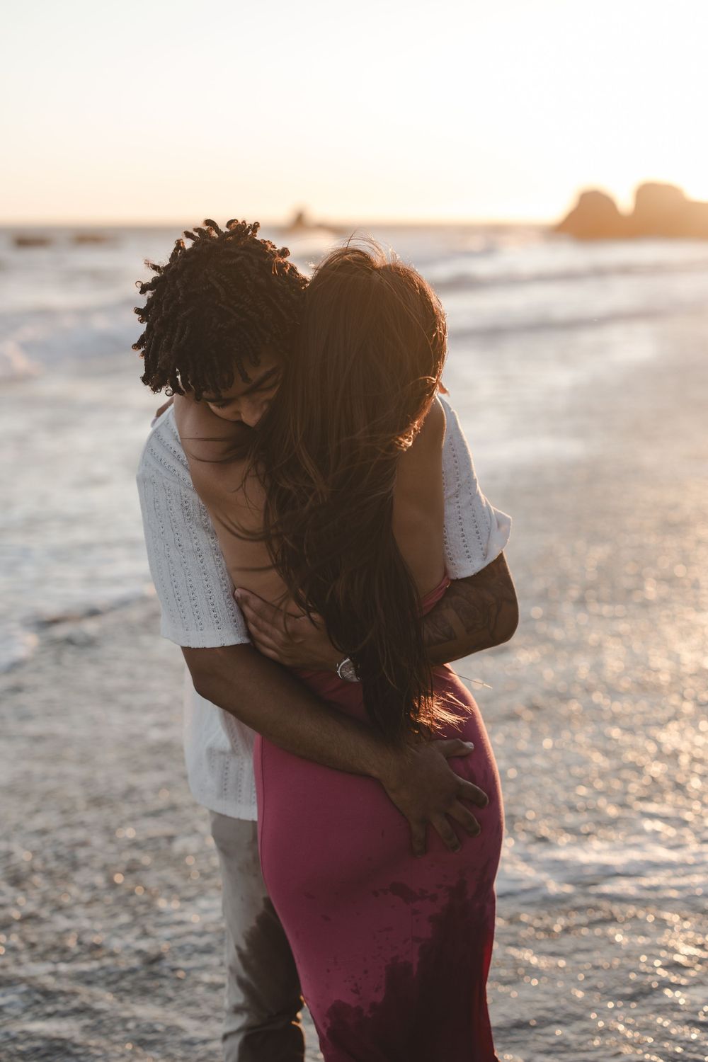 A couple embraces tenderly on a beach at golden hour with ocean waves in the background.
