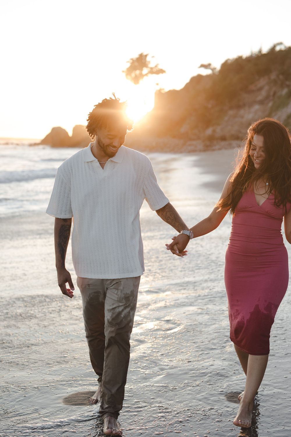 A couple walks hand in hand along a beach at sunset with golden light streaming through coastal cliffs.