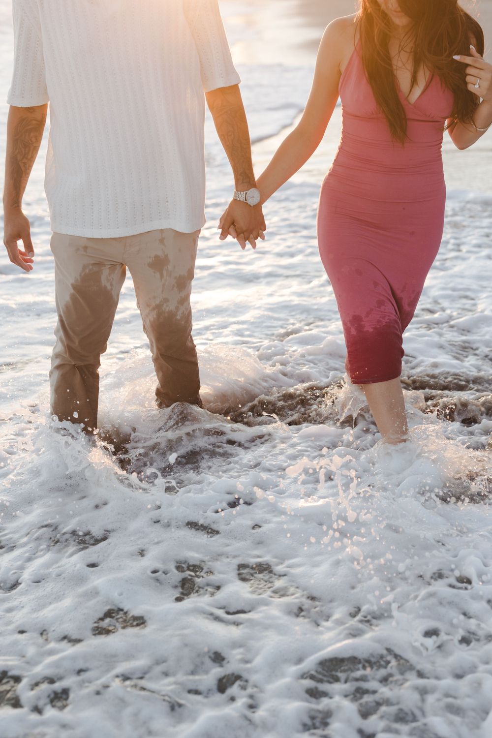 Couple holding hands while walking through ocean waves, wearing white and pink outfits.