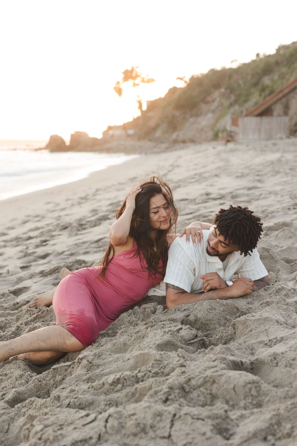 A couple lying on sandy beach at sunset sharing an intimate moment while waves crash nearby.