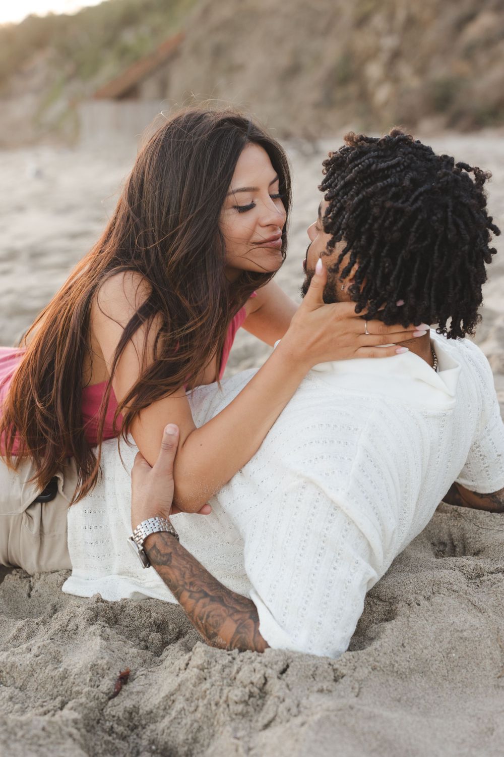 A romantic couple embraces intimately while lying on sandy beach during a golden hour photoshoot.