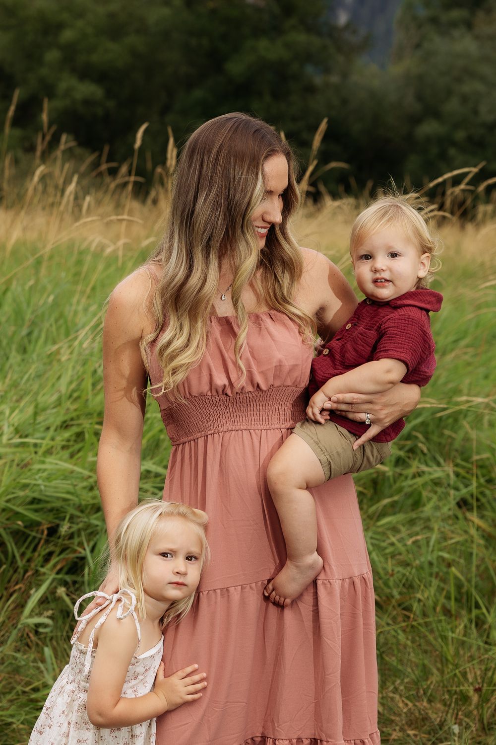 A mother in a pink dress holds a toddler in red while a young blonde girl hugs her side in a grassy field.