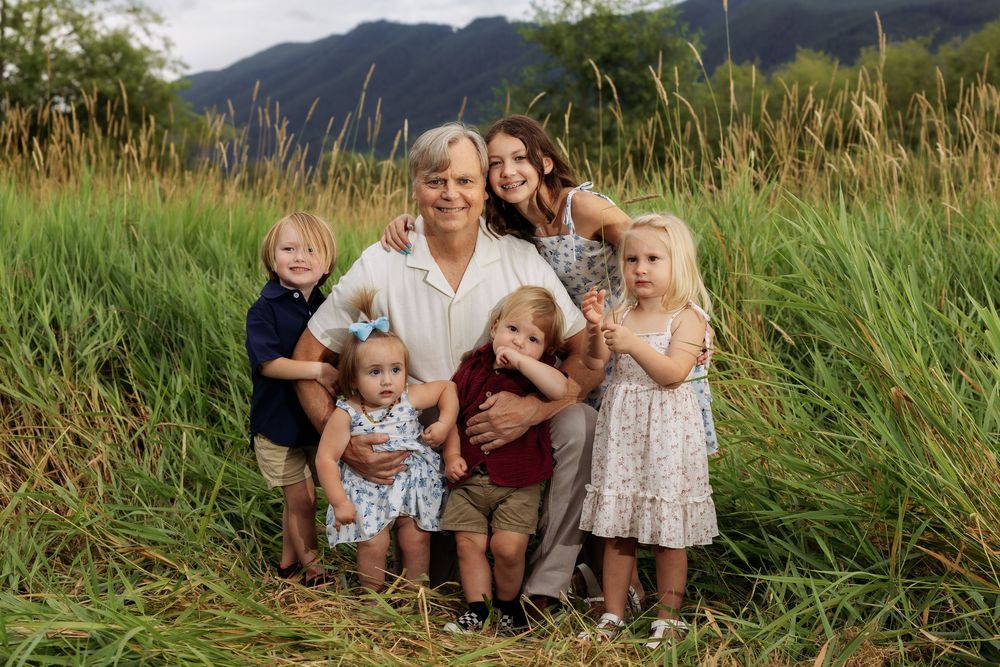 Family portrait with two adults and four young children posing in a lush green field with mountains in background.