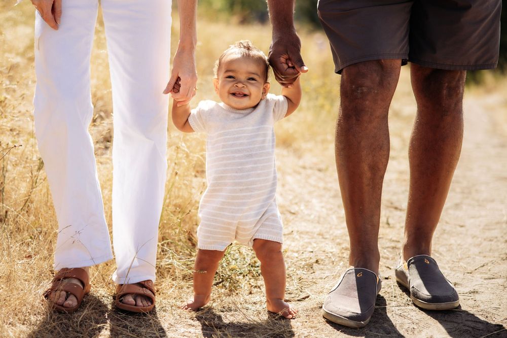 A smiling baby takes first steps outdoors, held by two adults on a sunny dirt path.