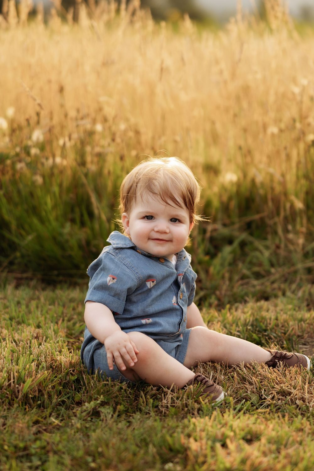 Adorable baby boy in blue romper sitting on green grass in front of golden wheat field, smiling at camera.