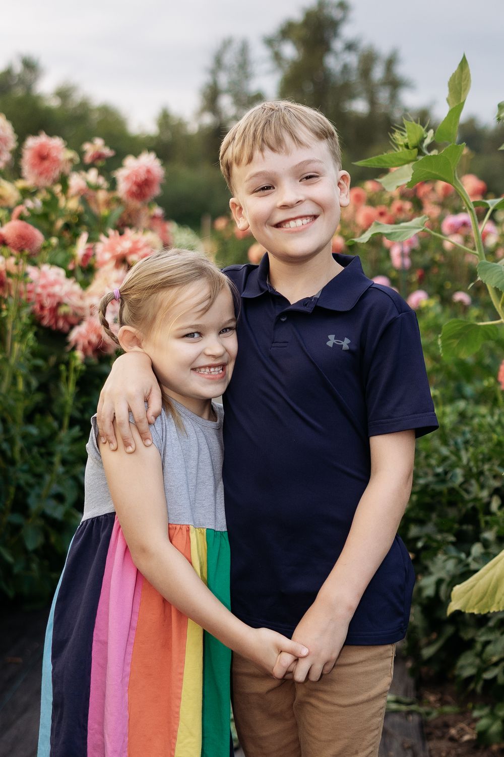 Two smiling children, a girl in a rainbow dress and a boy in a navy polo, hug in a garden with pink dahlias.