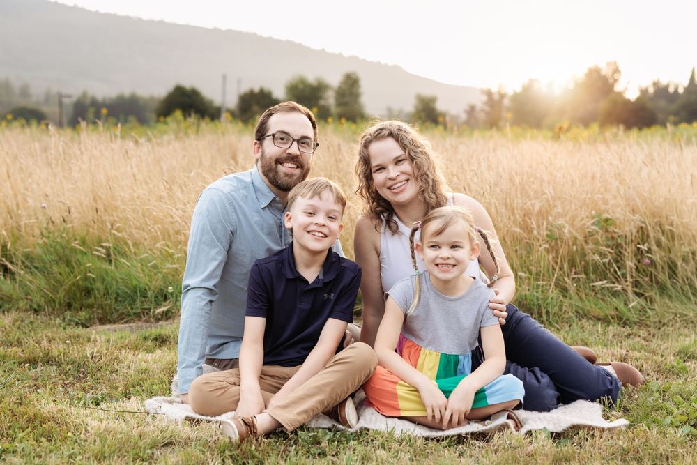 Happy family of four posing in a sunlit grassy field during a warm golden hour portrait session.
