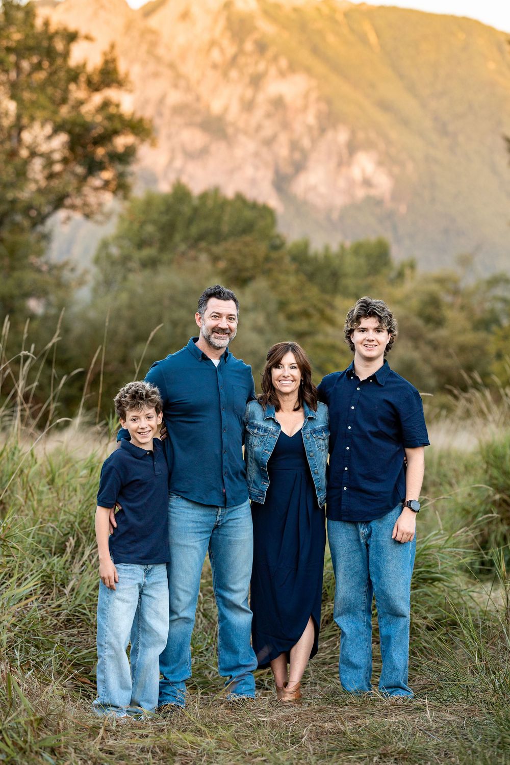 Family of four posing outdoors in coordinating navy and denim outfits with mountain and greenery backdrop.