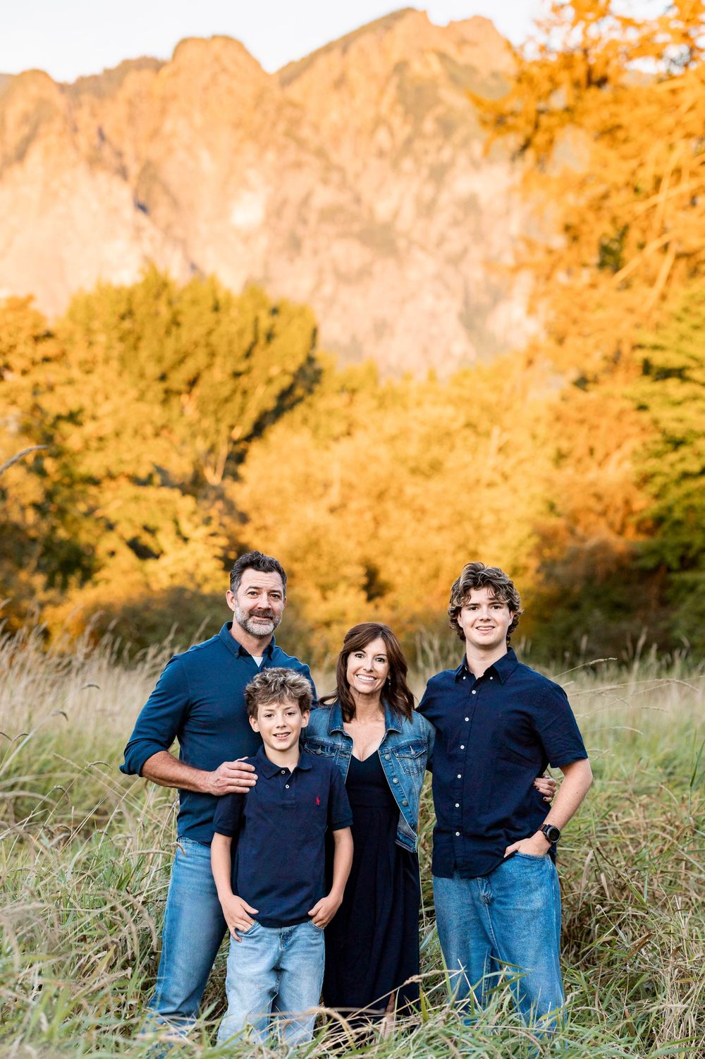 Family of four posing outdoors in fall foliage with mountain backdrop, wearing coordinated navy and denim outfits.