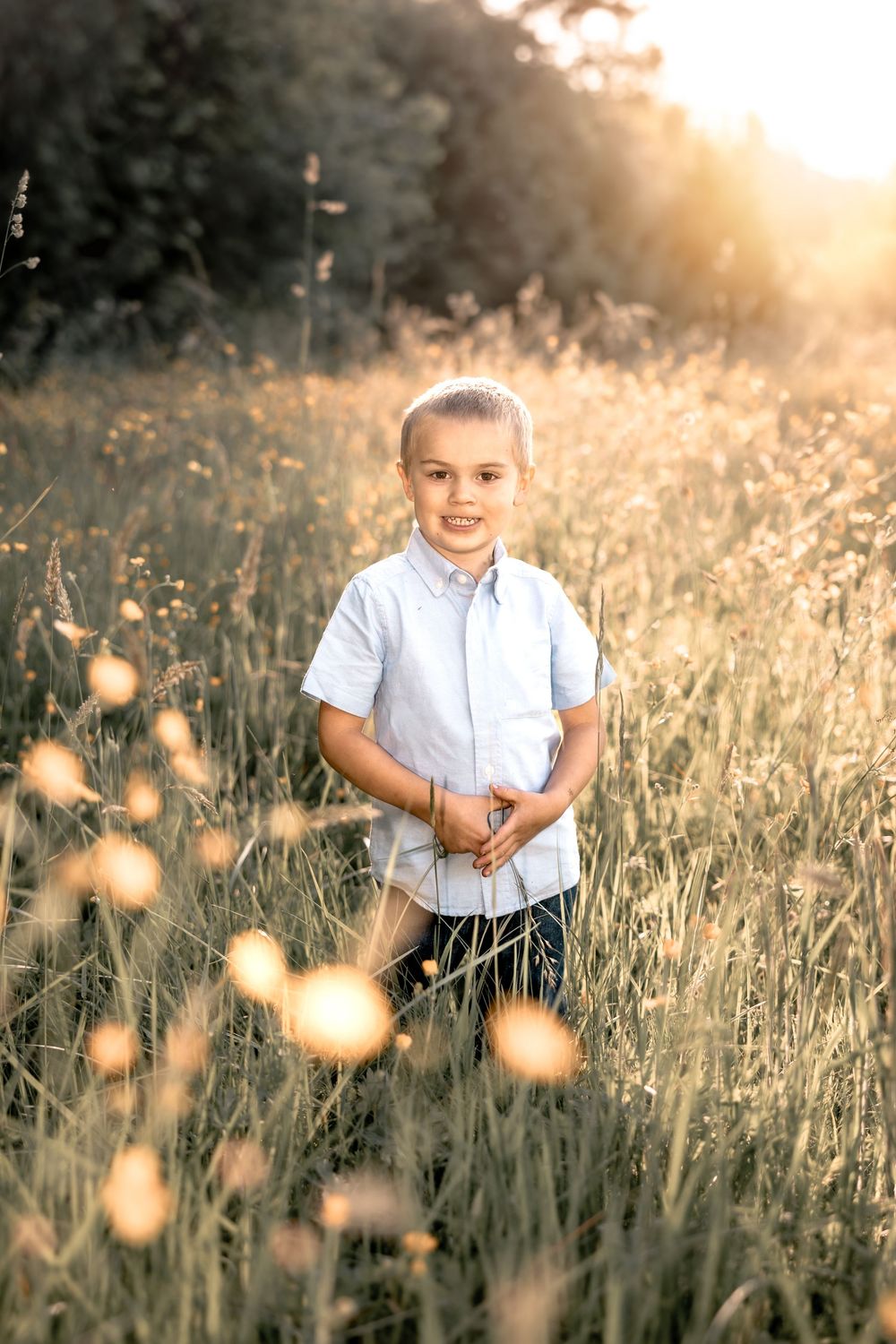 Young boy smiling in golden sunlit meadow with tall grass and wildflowers during sunset.