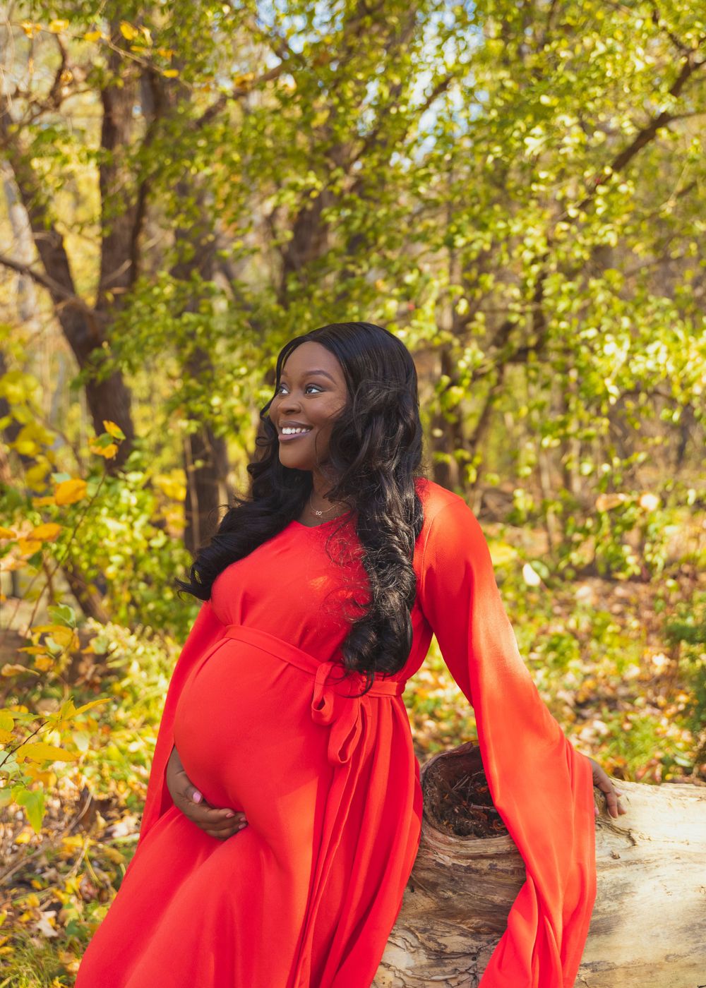 Outdoor maternity photo in vibrant red dress against autumn foliage background.