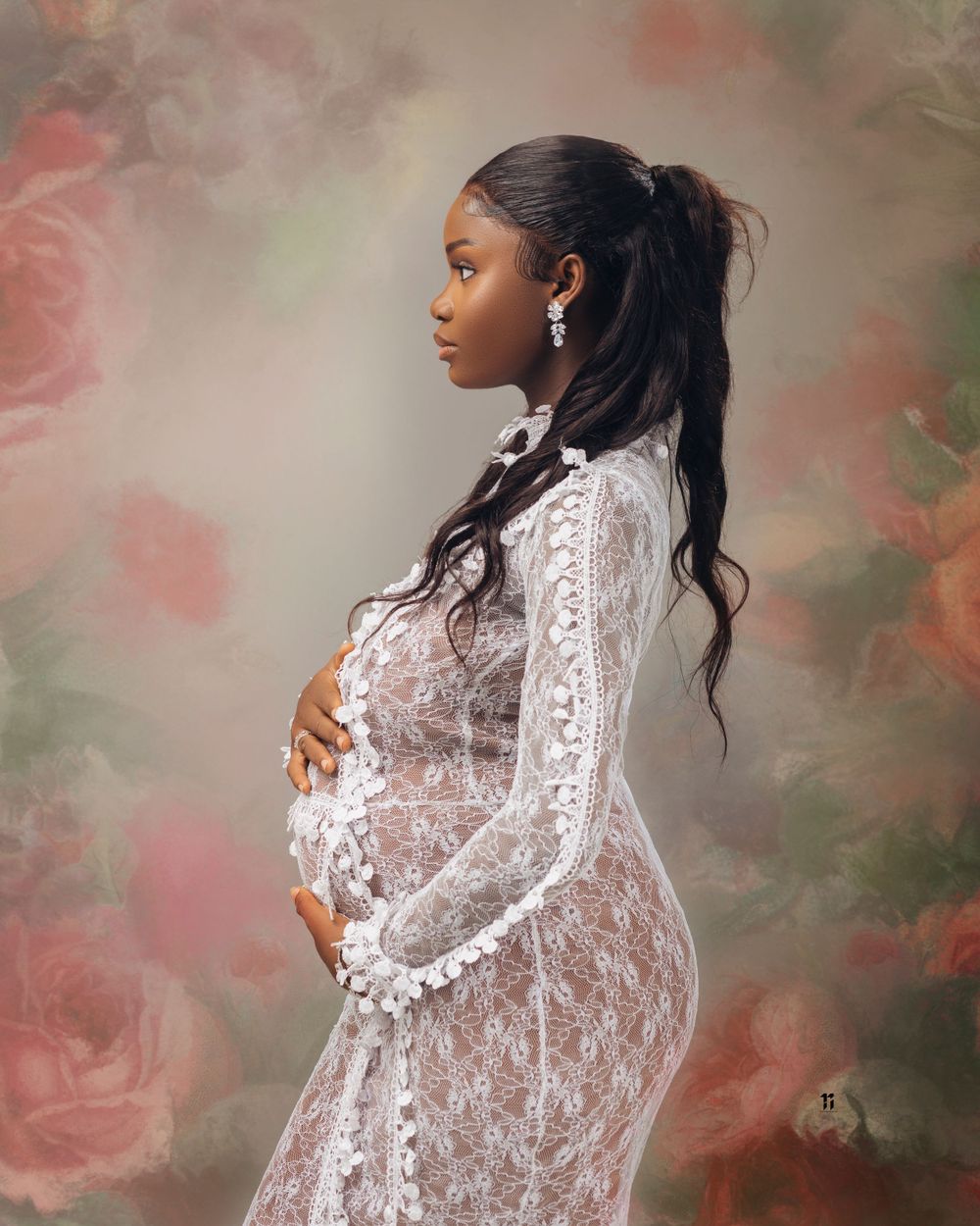 Maternity portrait in white lace dress with floral backdrop and soft lighting.