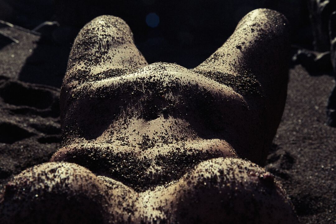 Artistic nude body covered in dark sand photographed from above on a shadowy beach.